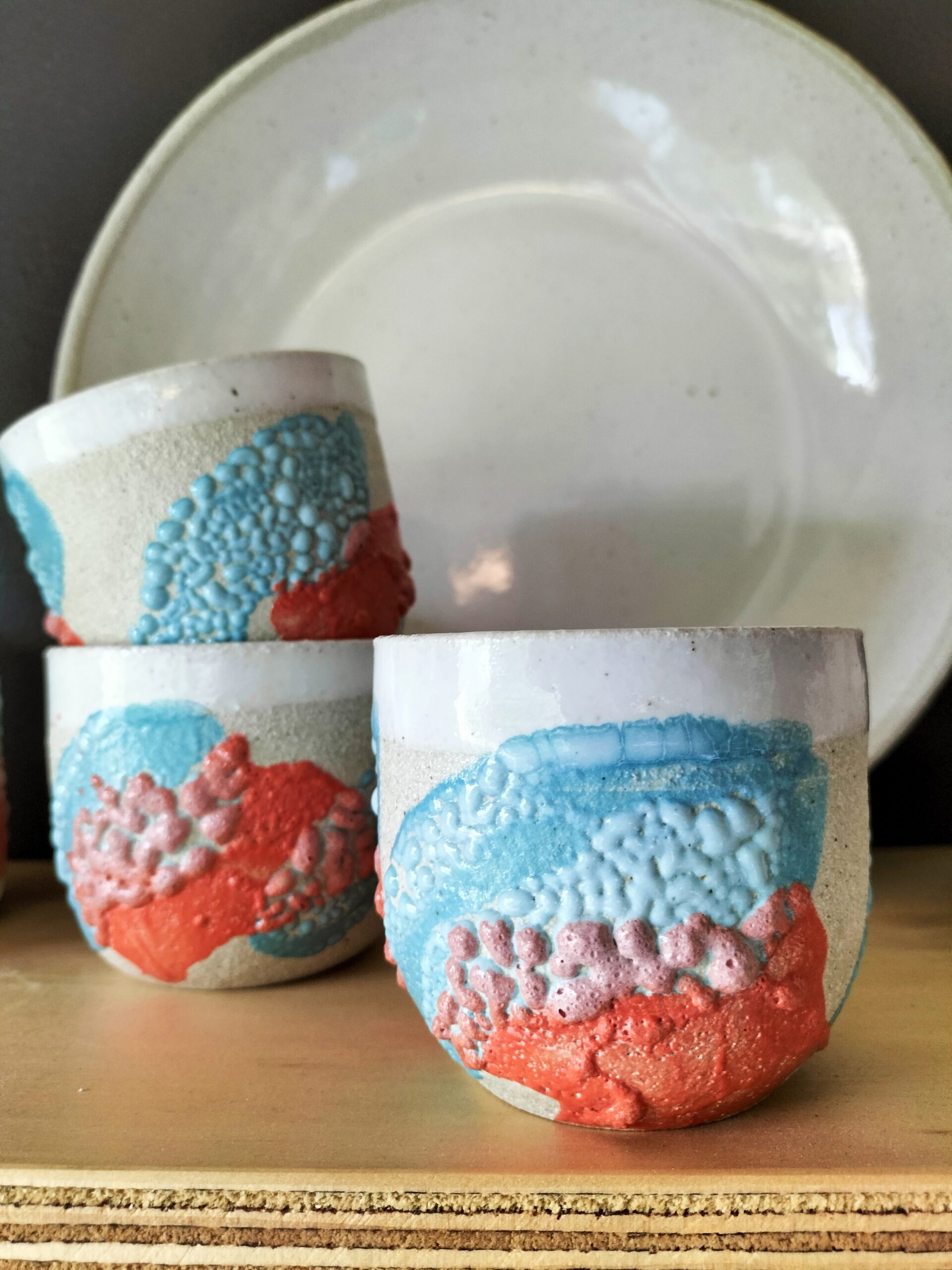 Image shows three cups in the blue and orange glaze called Bay of Fires and a white plate by Christie Lange in her Artist Profile Art Trails Tasmania